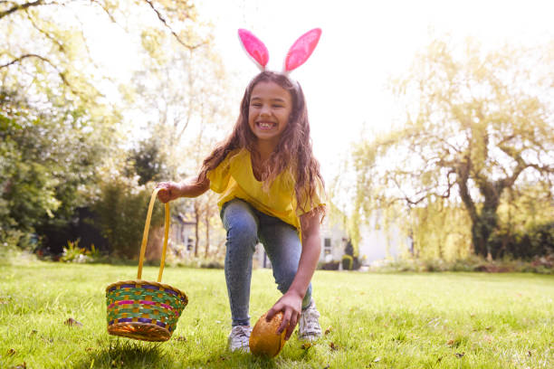 庭でイースターエッグハントでチョコレートエッグを見つけるバニー耳を身に着けている女の子の肖像画 - easter egg hunt ストックフォトと画像