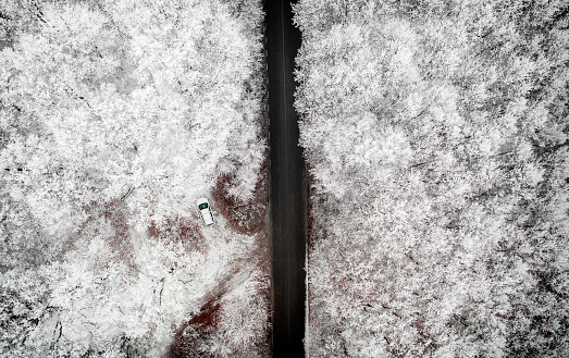 High angle color image, taken by drone, depicting a long straight road surrounded by dense forest covered with snow in the middle of winter. Parked in a clearing in the forest is a white car. The road runs directly through the centre of the image, with the frozen canopy of the forest either side of the road.