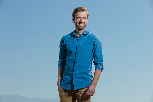 young casual man wearing blue shirt standing with one hand in pocket and looking away happy outdoor on blue sky