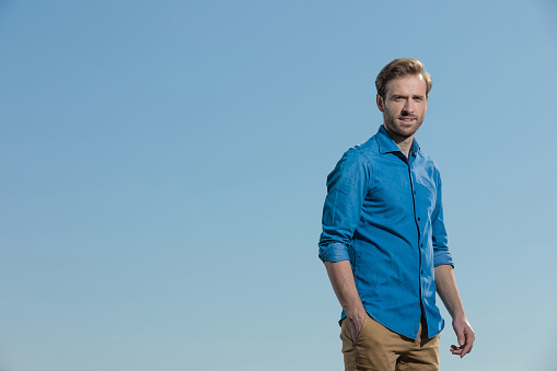 gorgeous casual man wearing blue shirt standing with hand in pocket and looking at camera relaxed outside on blue sky