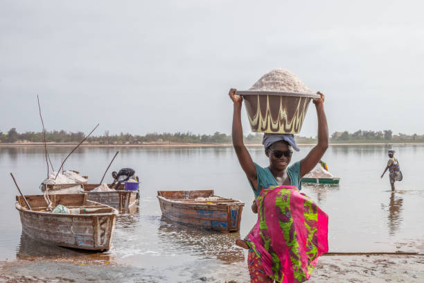 Dakar, Senegal - june 28, 2019: Young woman unloading salt in a bucket over her head with the salt-extracting boats in the background of the image