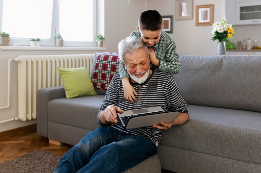 Grandfather and grandson using laptop together and laugh while looking at something funny on a laptop. Family relationship with grandfather and grandson. Grandpa and grandchild using technology at home in the livingroom.