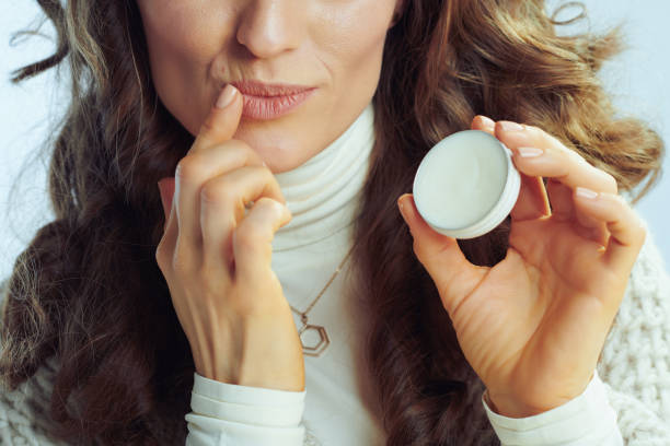 Closeup on modern housewife applying lip balm Closeup on modern housewife with long wavy hair in neck sweater and cardigan applying lip balm on winter light blue background. beeswax photos stock pictures, royalty-free photos & images