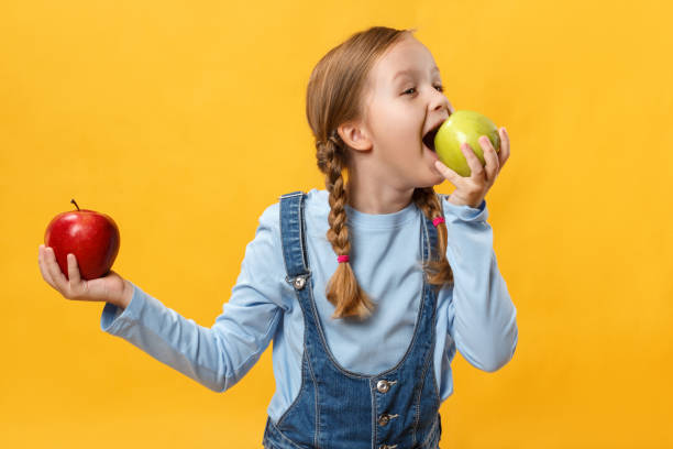 концепция здорового питания. ребенок кусает яблоко. маленькая девочка на желтом фоне - apple eating little girls green стоковые фото и изображения