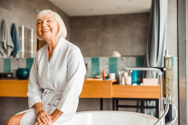 Cheerful elderly woman relaxing in spa salon Smiling senior lady in bathrobe sitting on the edge of bathtub stock photo bathrobe stock pictures, royalty-free photos & images