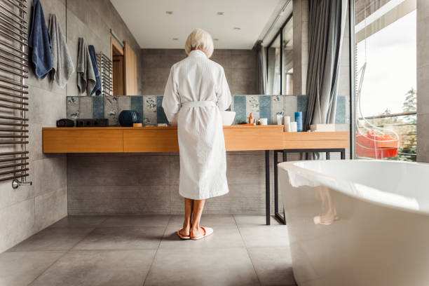 Old woman standing in front of big mirror Elderly lady in white bathrobe standing by the sink in bathroom stock photo bathrobe photos stock pictures, royalty-free photos & images