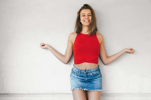 Beautiful teen girl in short red top and denim mini skirt laughing against a concrete wall with open arms