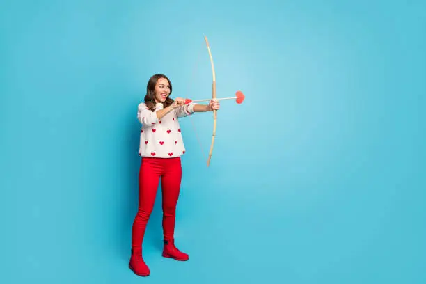 Full length body size view of nice attractive charming fashionable cheerful cheery girl wearing, festal theme party clothes shooting arrow isolated on bright vivid shine vibrant blue color background
