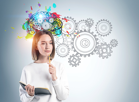 Thoughtful young woman in casual clothes with notebook standing near gray wall with colorful brain sketch with gears drawn on it. Concept of brainstorming