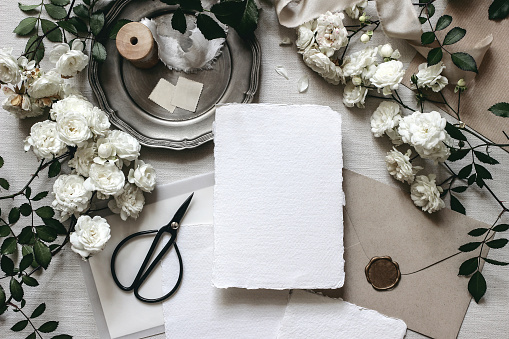 Moody wedding table mockup scene. Stationery composition with fading white rose flowers, silver plate, black scissors, envelopes and blank greeting cards. Grey textured background, flat lay, top view.