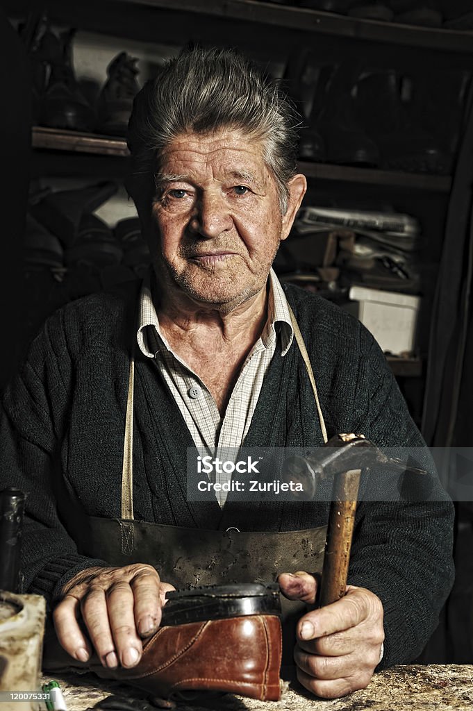 Old man, shoemaker, repairing  handmade shoe in his workshop Old man, shoemaker, repairing old shoe in his workshop and looking at the camera Active Seniors Stock Photo