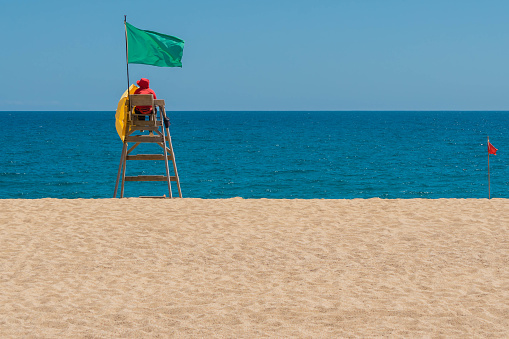 Lifeguard on rescue post at sandy beach near sea with green flag. Lifeguard searching sea areal. Safe vacation concept. Clean endless beaches. Copy space.
