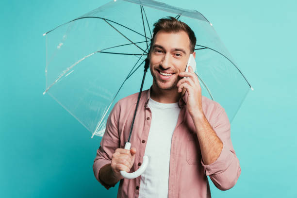 istock cheerful man holding umbrella and talking on smartphone, isolated on blue 1200755709