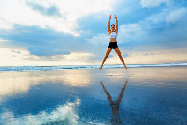 Young woman doing jumping jack or star jumps exercise to burn fat, keep fit. Sunset beach, blue sky background. Healthy lifestyle at training camp, outdoor fitness activity, family summer holiday.