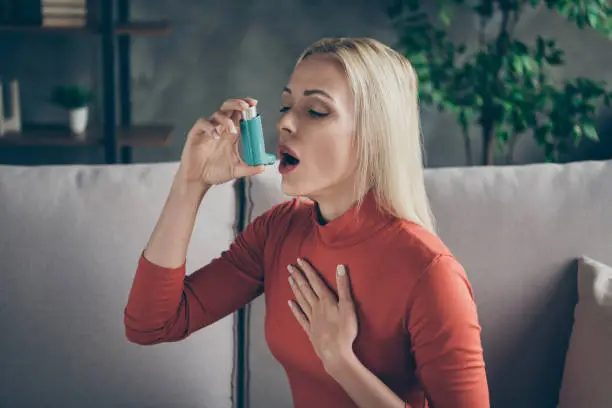 Photo of Profile photo of unhealthy pretty blond lady suffering allergy holding inhaler breathing in feel better sitting couch wear orange pullover shirt living room indoors