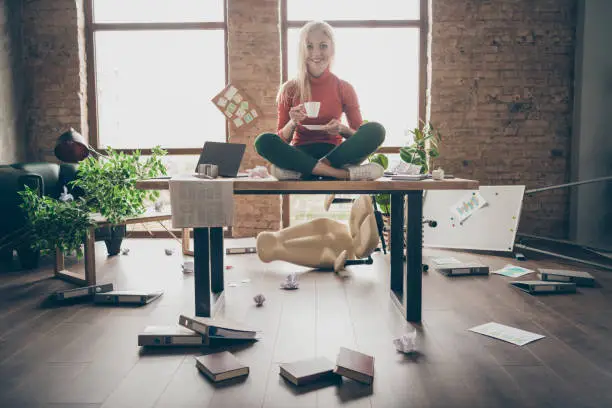 Full body photo of happy trader woman sit on table crossed legs feel carefree, careless rest relax hold white coffee cup in messy office loft