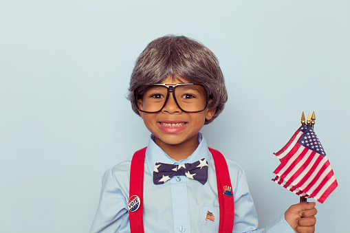 A young boy dressed as a United States of America campaign worker wearing a bowtie, wig, eyeglasses, and suspenders smiles at the camera as he is excited for the upcoming election season. He enjoys the right to vote and to have his voice heard in politics.