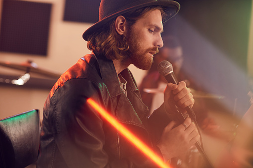 Side view portrait of handsome bearded man singing to microphone during music concert with contemporary band in studio, copy space