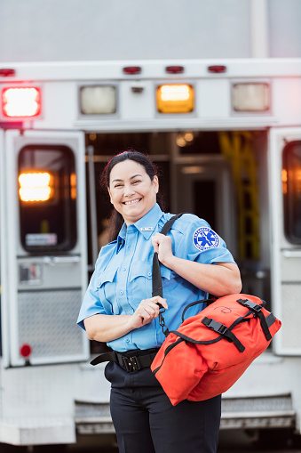 A mature Hispanic woman in her 40s, a paramedic, standing at the rear of an ambulance, by the open doors. She is looking at the camera with a confident expression, smiling, carrying a medical trauma bag on her shoulder.