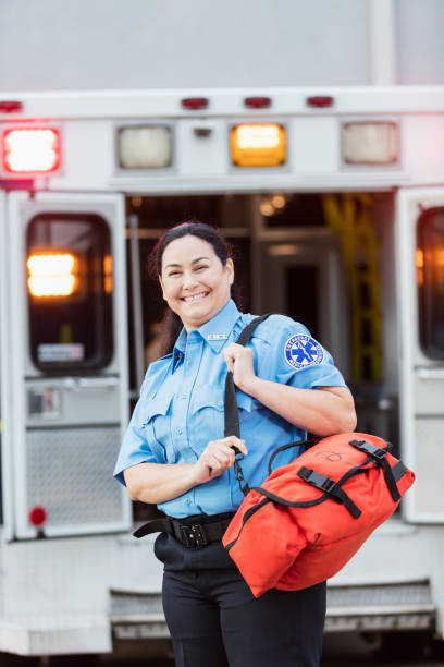 Female paramedic standing at the rear doors of an ambulance A mature Hispanic woman in her 40s, a paramedic, standing at the rear of an ambulance, by the open doors. She is looking at the camera with a confident expression, smiling, carrying a medical trauma bag on her shoulder. paramedic photos stock pictures, royalty-free photos & images