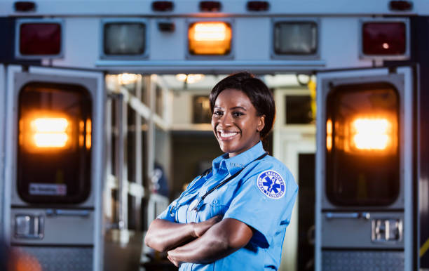 African-American woman working as paramedic A young African-American woman standing, arms folded, smiling at the camera. The back of an ambulance with doors open and emergency lights on, is out of focus behind her. She is working as an EMT or paramedic. ambulance photos stock pictures, royalty-free photos & images