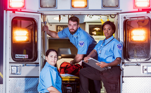 A multi-ethnic group of three paramedics at the rear of an ambulance, ready to climb in through the open doors.  The two women their male colleague have serious expressions.