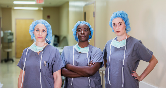 A multi-ethnic group of three female doctors or nurses standing in a hospital corridor, wearing scrubs and surgical caps. The team of healthcare works are staring at the camera with serious expressions.