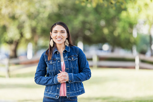 A young Hispanic woman in her 20s standing outdoors at a park, smiling at the camera. She is wearing a denim jacket and jeans.