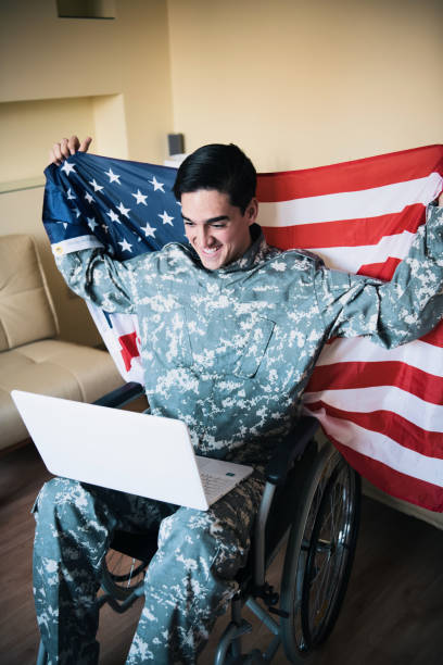 istock Retired Soldier With Physical Disabilities is Using a Laptop and Holding US Flag 1200817702