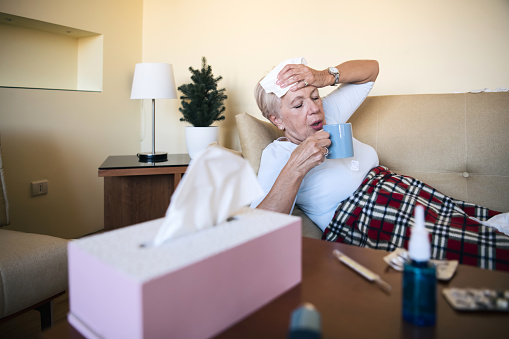 An Older sick woman with temperature is lying in her bed covered with a blanket. A senior woman is sweating due to the flu and taking a hot cup of tea.