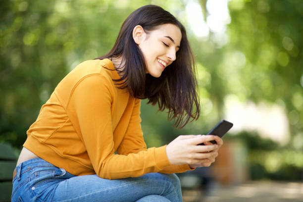 young woman smiling and looking at mobile phone while sitting on park bench