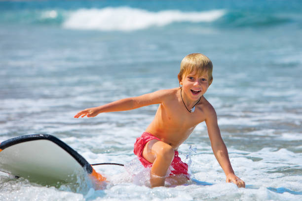Happy boy - young surfer learning ride, jump from surfboard with fun, run to beach. Active family lifestyle. Kids surf lessons, outdoor water sport activity in surfing camp. Summer vacation with child
