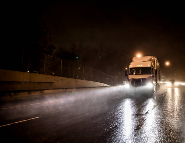 Big rig white semi truck transporting cargo in semitrailer driving on the night wet road with heavy rain and water dust Big powerful bonnet rig industrial grade semi truck with loaded semi trailer transporting commercial cargo running at night time with turned on headlights on the wet raining road with reflection truck rain stock pictures, royalty-free photos & images