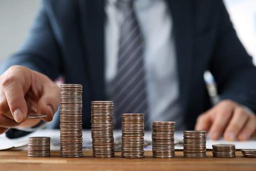 Close-up view of businessman in suit increasing budget by putting penny on top. Coins in ascending order. Selective focus on cash. Financial growth and revenue concept