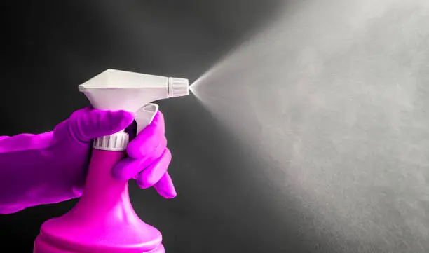 Woman wearing pink rubber gloves using pink spray bottle and spraying liquid mist in air, cool lighting effect. Lot of copy space.
