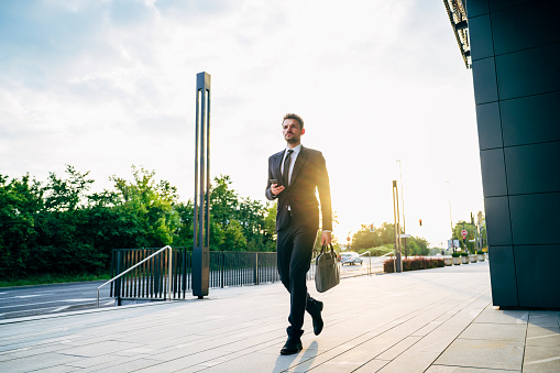 Low angle view of approaching Ljubljana businessman in early 30s holding smart phone and briefcase while arriving for work in morning.