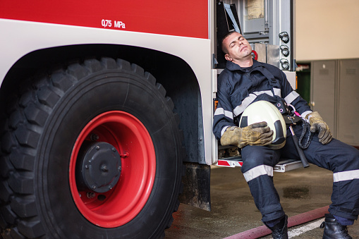 Exhausted male firefighter sitting on the fire truck with closed eyes and taking a short rest