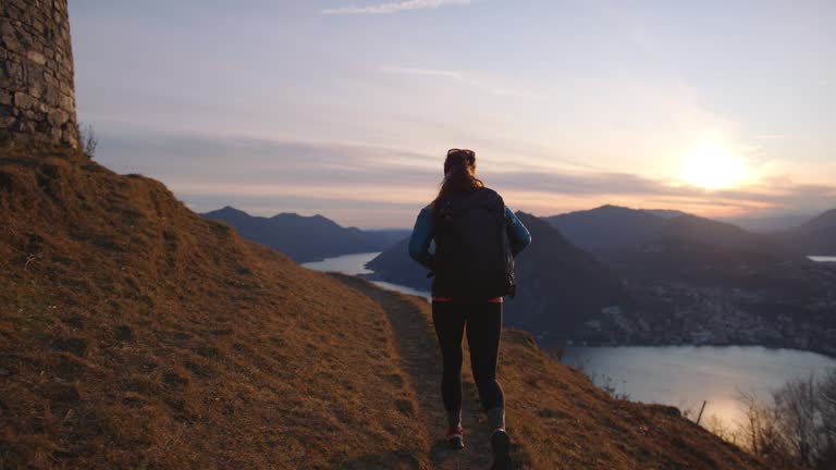 Female hiker follows trail to mountain top with view of city, lake and sunset below