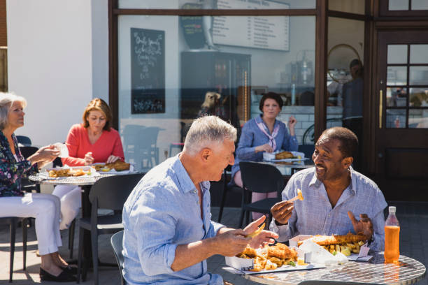 Senior male friends enjoying fish and chips outside cafe Cheerful male friends eating lunch together on sunny day, seaside, friendship, enjoyment outdoor dining photos stock pictures, royalty-free photos & images