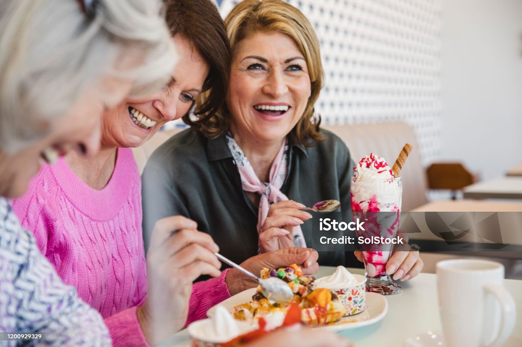 Cheerful senior women laughing together in ice cream parlour Happy female friends on holiday together eating ice cream sundaes, fun, carefree, laughing, bonding Eating Stock Photo