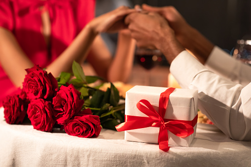 Happy Anniversary. Romantic Background Of Couple Holding Hands. Wrapped Gift And Red Roses Lying On Served Restaurant Table. Selective Focus