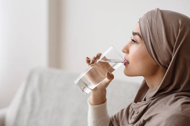 durstige muslimische frau in kopftuch trinken mineralwasser aus glas - zurückhaltende kleidung stock-fotos und bilder