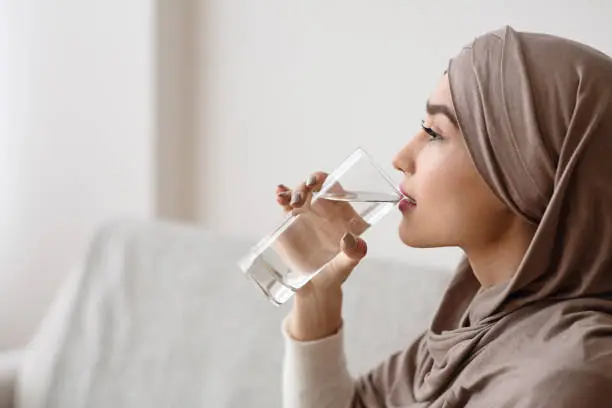 Thirsty islamic woman in headscarf drinking mineral water from glass at home, selective focus, empty space