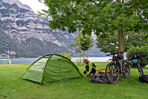 Сyclist tourist puts up tent in campsite by lake. Cloudy day. Walensee, Switzerland, Europe.