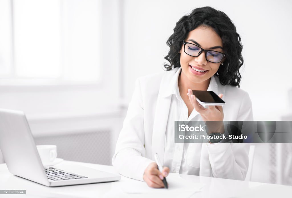 Smiling hispanic woman using voice assistant on phone Voice Assistant Concept. Mexican manager recording message on smartphone, writing notes at office, empty space Dictation Stock Photo