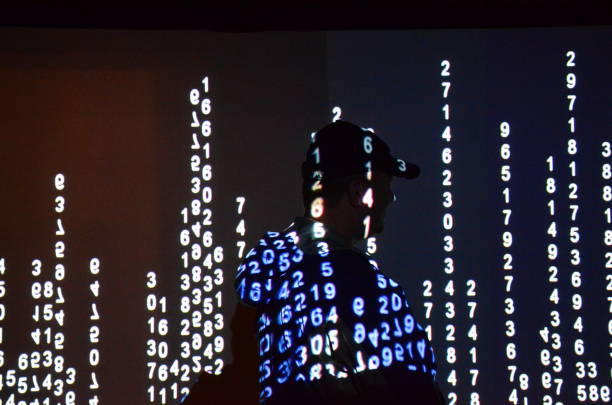 A man stands in front of a wall that is illuminated with numbers in a row, which are being shown against a black wall. It gives an interesting effect.
