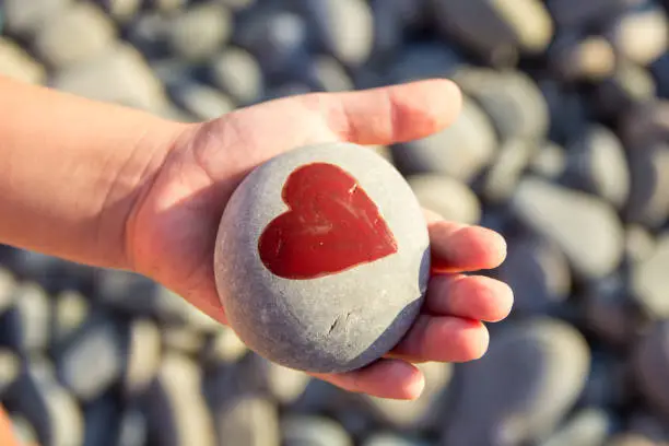 Photo of pebbles with a painted heart in the hands of a child on the background of a pebble beach