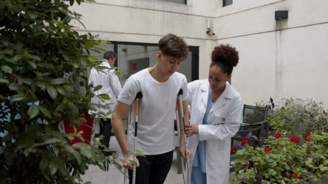 Female black therapist helping teenage male patient while he walks with crutches at the patio of physical rehab clinic