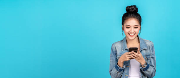 bannière de femme asiatique heureuse se sentant le bonheur et le smartphone de dactylographie restant sur le fond bleu. fille mignonne d’asia souriant utilisant la chemise occasionnelle de jeans et connectent l’achat en ligne d’internet et surfer. - mannequin métier photos et images de collection