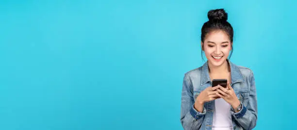 Photo of Banner of Happy asian woman feeling happiness and standing typing smartphone on blue background. Cute asia girl smiling wearing casual jeans shirt and connect internet shopping online and surfing.